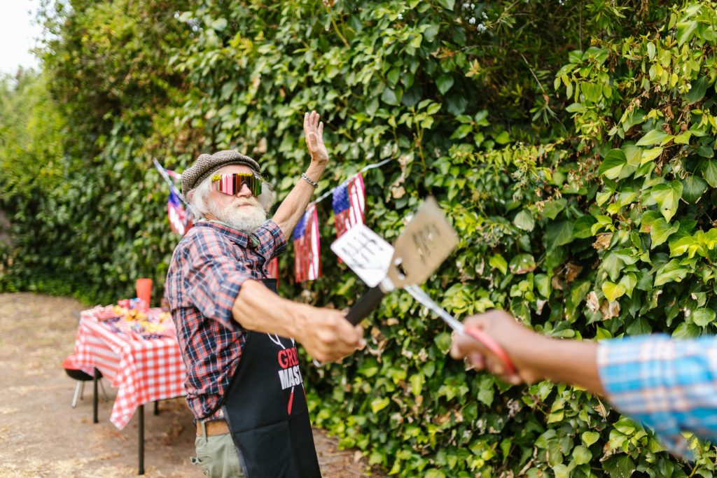 Home Elderly man enjoying a lively backyard barbecue with American flags and grill tools.